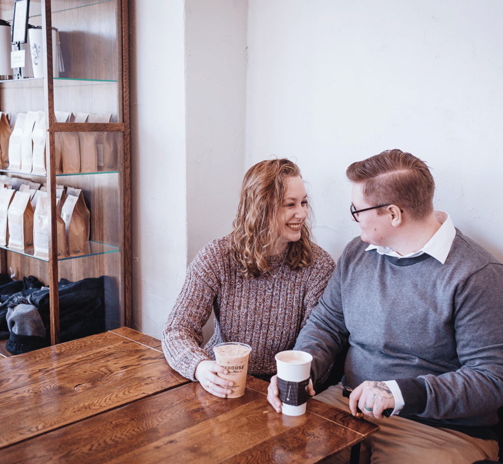 Image of a couple looking at each other smiling in a coffee shop. The couple each has a coffee cup in their hands. 