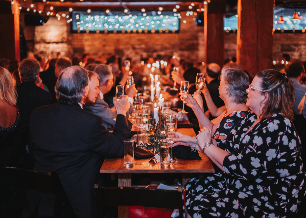 Image of people sitting at a long rectangle table with their glasses raised for cheers for wedding speeches. This image shows the romantic wedding venues of Minnesota. 