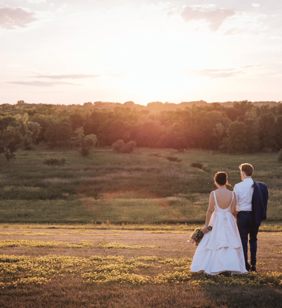 Image of a newly married couple standing with their backs to the camera in an open grass feild as the sun is setting infront of them. 