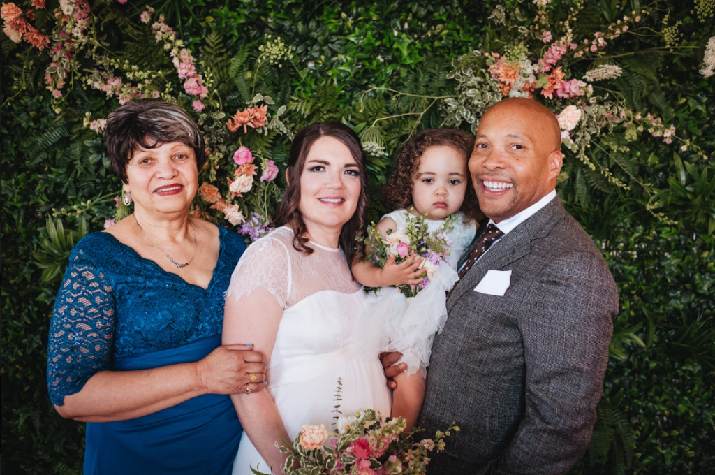 Image of a couple smiling infront of a green wall with flowers behind. There is a bride, groom, child and mother. 