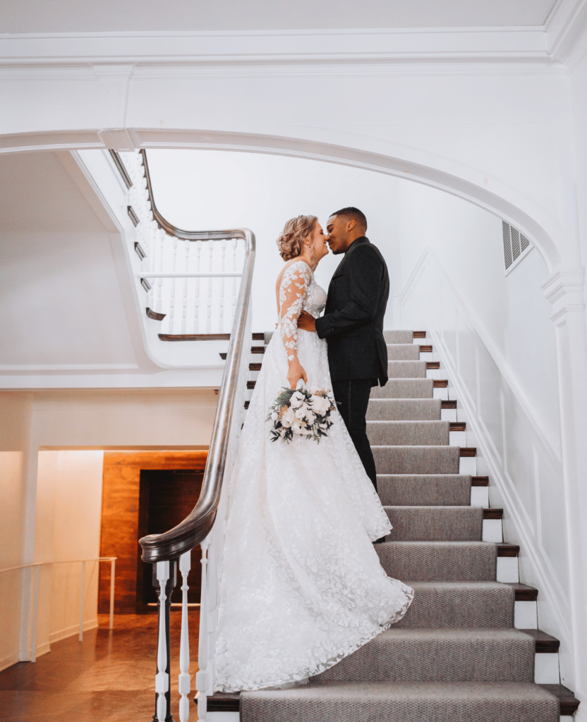 Image of a bride and groom halway up a staircase kissing. The walls are white and the staircase has a grey runner. 