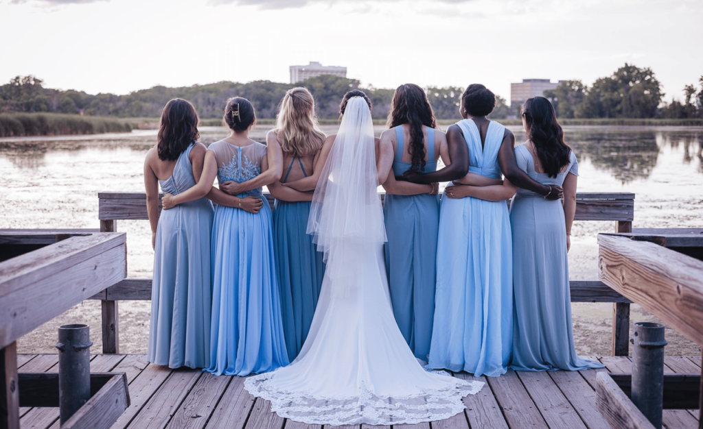 Image of bridal party standing side by side with arms linking staring away from camera. Bride is in the middle of the photo, they are standing on a dock. 