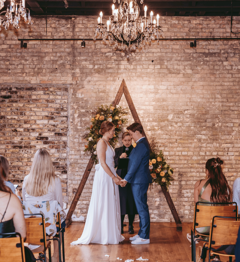 Image of a couple standing getting married infront of friends and family, there is a brick wall behind them and a wooden frame with flowers. 