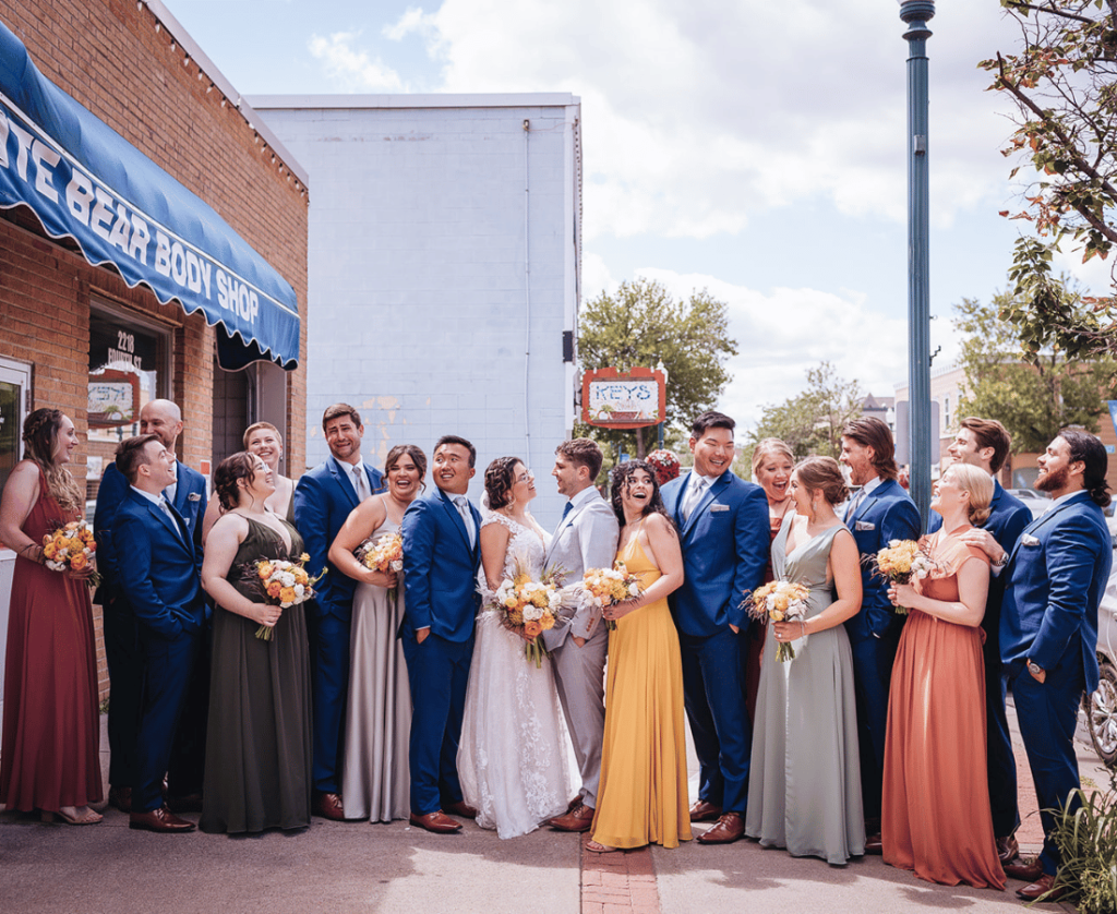 Image of wedding party photos being taking with the large wedding party laughing and interacting with each other. Bride and Groom are looking at each other. 