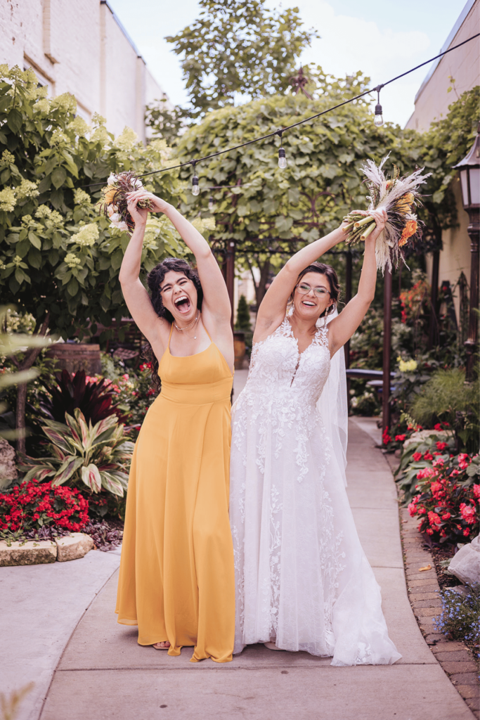 Image of a bride and maid of honour with their arms raised high with bouquets and bumping their hips together. 