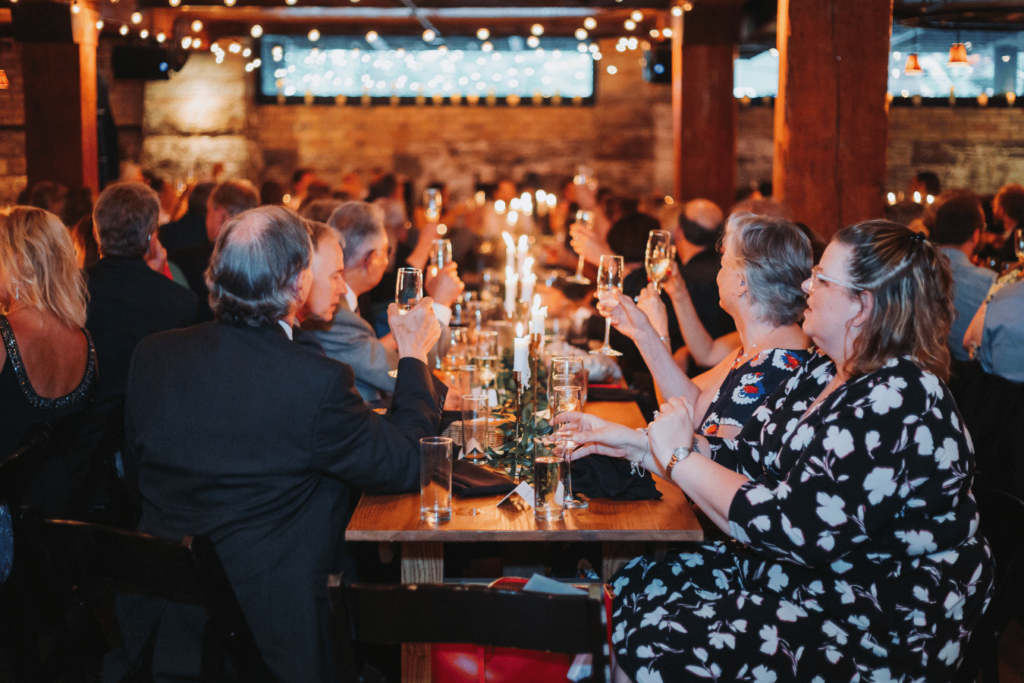 Image of wedding dinner reception with folks raising their glasses in cheering. This image is showing how to start wedding planning with thinking of your guest list and who you will invite. 