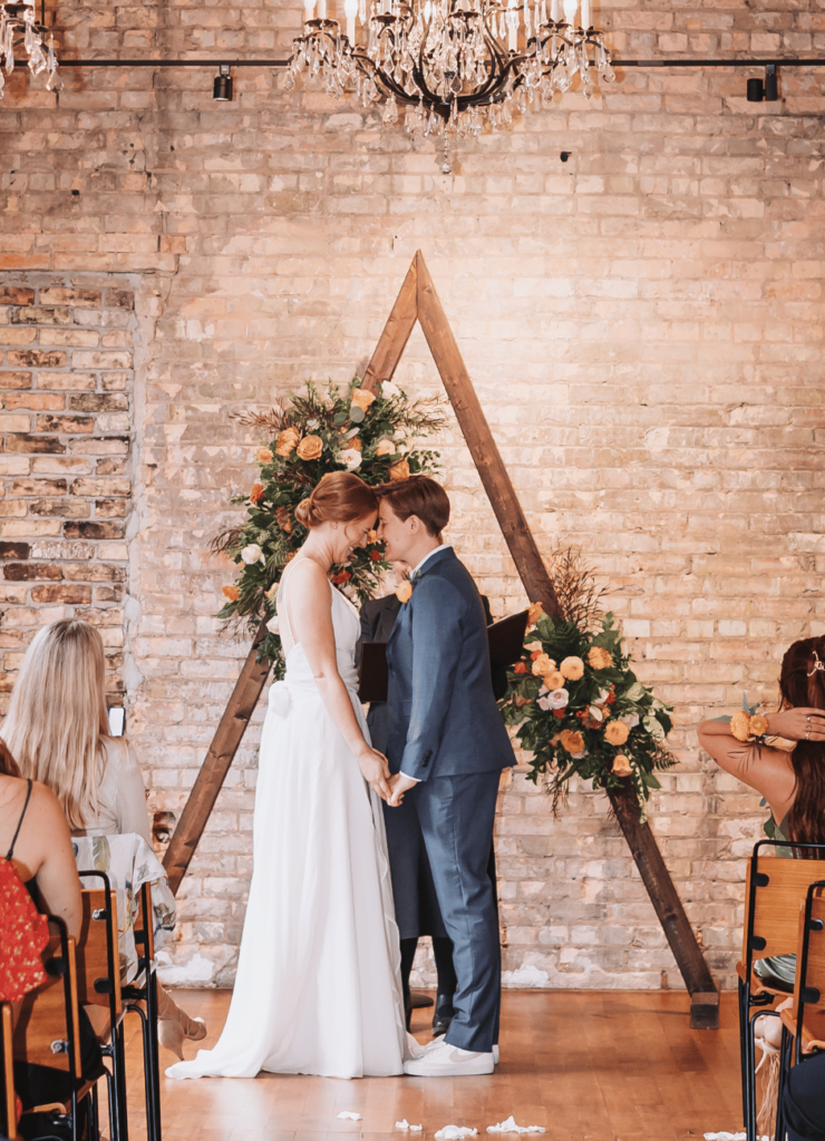 Image of a couple standing at the wedding alter during their ceremony. There is a wooden arch behind them.