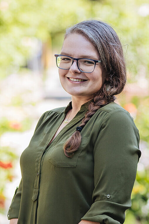 headshot of a creative business owner in the Twin Cities. She is wearing a green shirt and a braid in her hair and is surrounded by greenery and gardens.