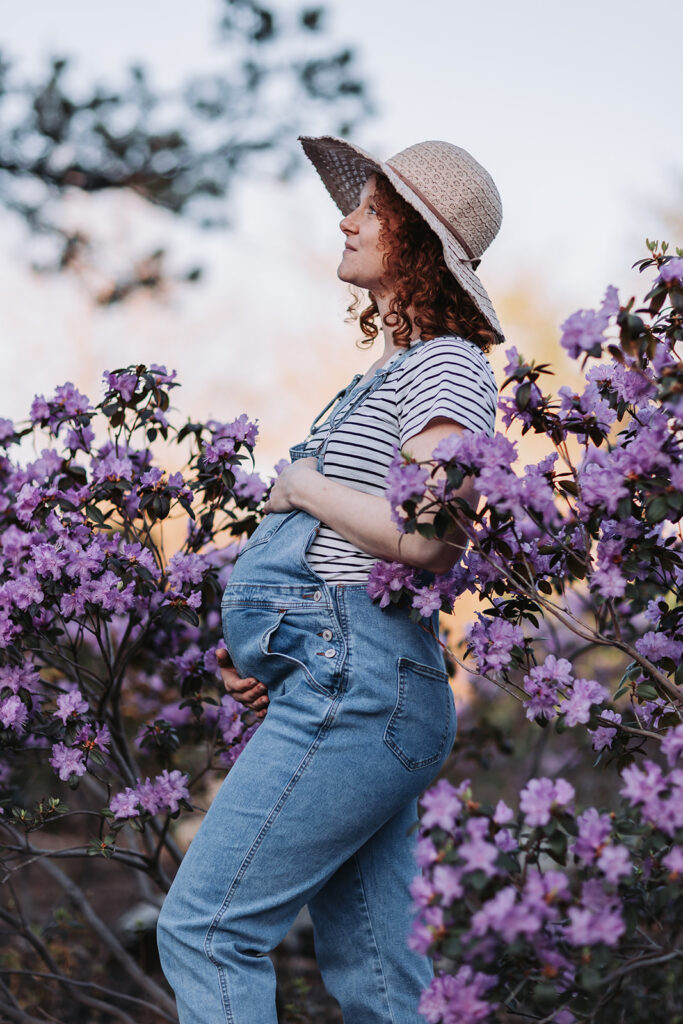 Pregnant woman in overalls poses amongst purple flowers for her maternity photos in Minneapolis