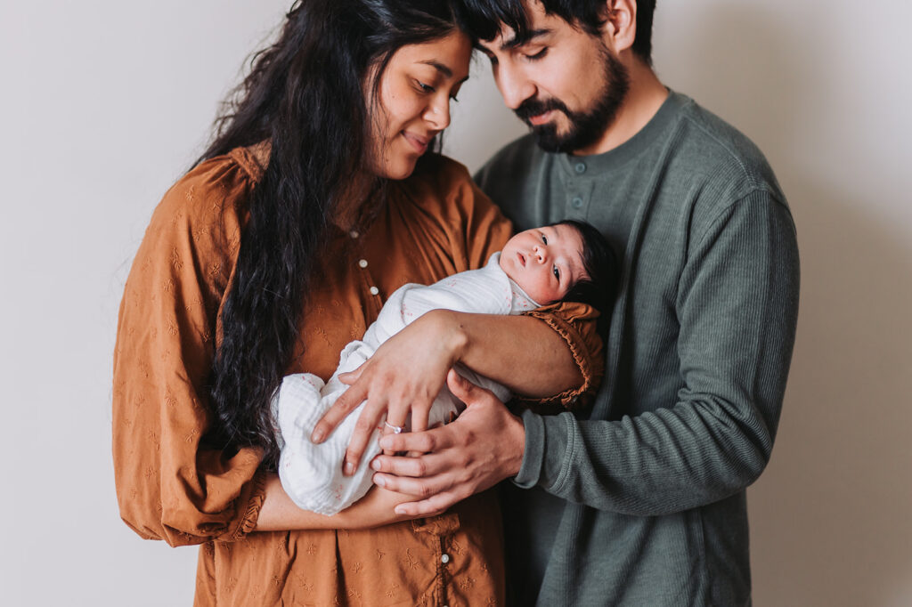 Couple stands against a white wall holding their newborn baby who looks at the camera during their newborn photo session