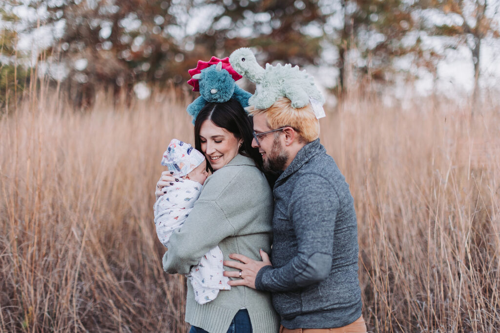 Funny newborn photo with parents with stuffed toys on their heads, holding their baby outdoors in a field